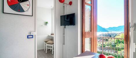 Et moderne værelse med fjernsyn, et indrammet abstrakt kunstværk, et spiseområde med bord og stole, samt en balkon med udsigt til bjergene.