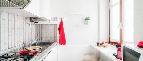 Cocina compacta con estufa a gas, lavamanos y microondas. Hay una toalla roja colgada en la pared, una pequeña planta en una repisa y una ventana que proporciona luz natural.