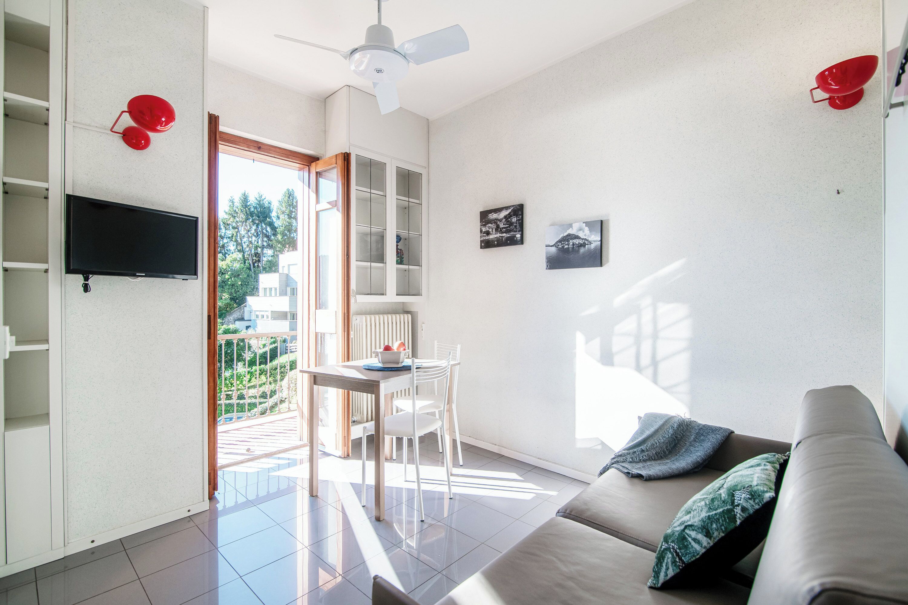 Una sala de estar moderna con un sofá, un televisor montado en la pared, una zona de comedor con mesa y sillas, y una vista al exterior a través de una puerta abierta.