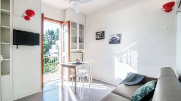 Una sala de estar moderna con un sofá, un televisor montado en la pared, una zona de comedor con mesa y sillas, y una vista al exterior a través de una puerta abierta.