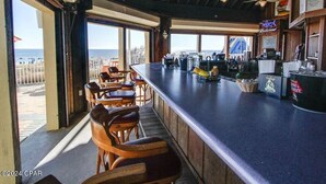 Eine Bar mit Holzhockern und einer Theke, großen Fenstern und Blick auf den Strand.