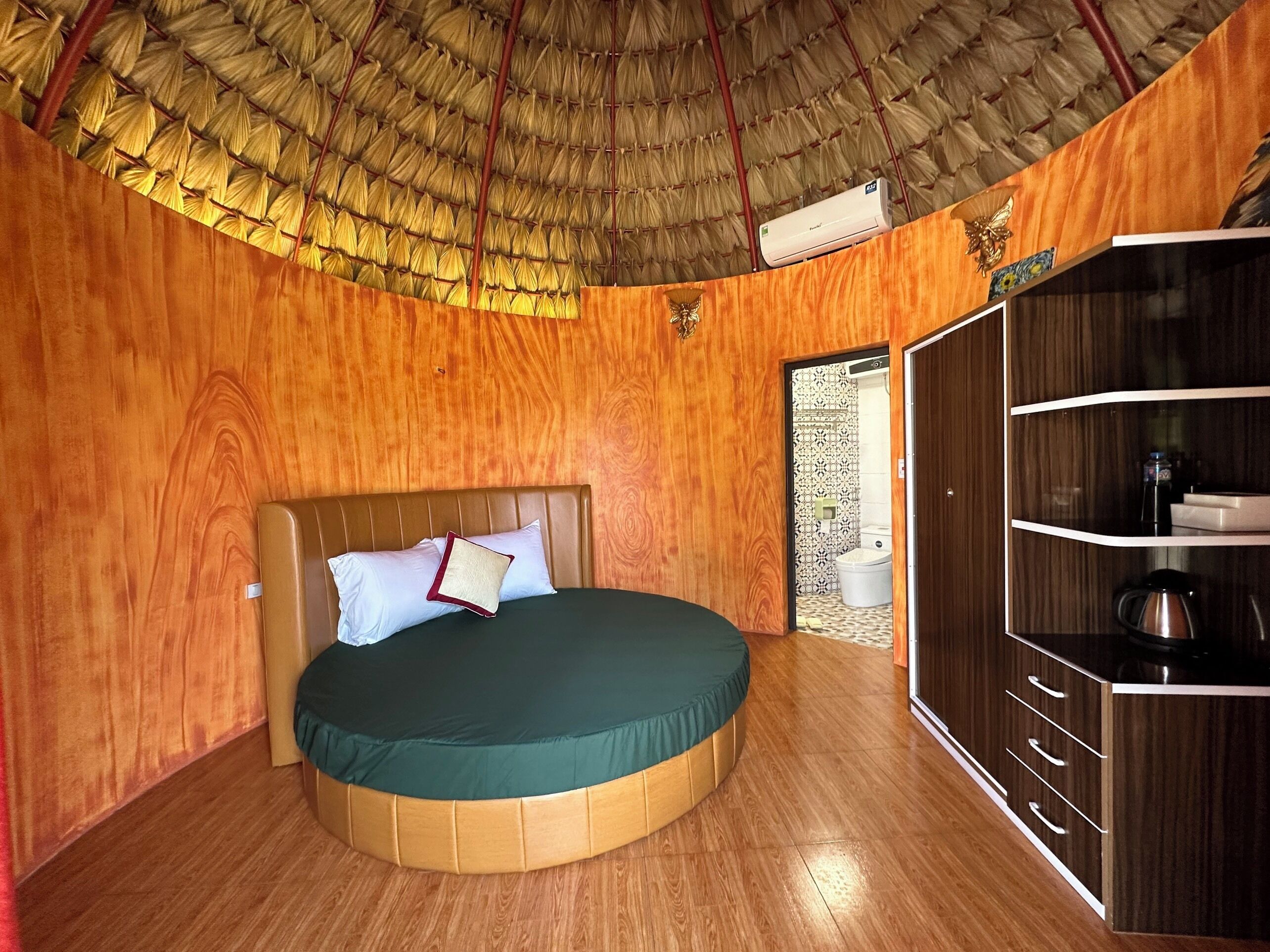 A room with a round bed, wooden walls, a thatched roof, and a small kitchenette.
