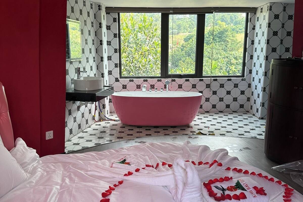 Un dormitorio con una cama grande, una bañera rosa y una ventana con vistas a la vegetación.