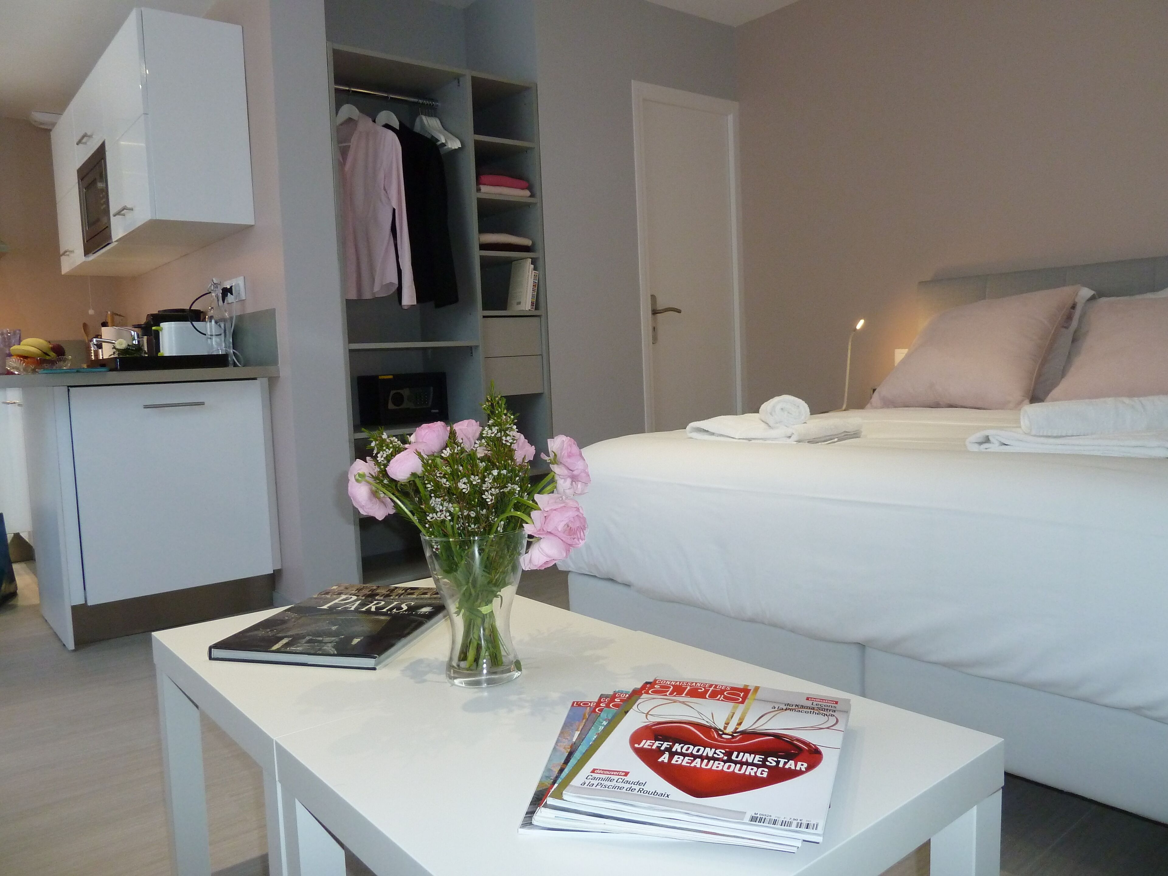 A hotel room with a bed, a bedside table with a magazine, a vase with flowers, a wardrobe, and a kitchenette.