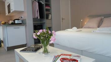 A hotel room with a bed, a bedside table with a magazine, a vase with flowers, a wardrobe, and a kitchenette.