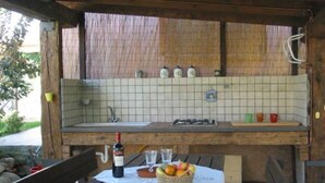 A rustic outdoor kitchen with a tiled backsplash, a wooden bench, a table with a wine bottle and fruit basket, and a hanging light fixture. - Rossini Apartment (Urbania)