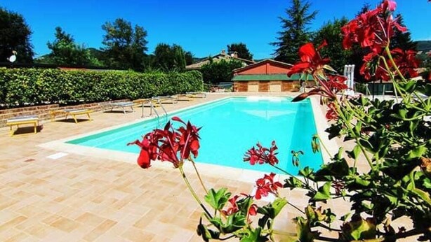 A swimming pool with a tiled deck, surrounded by greenery and a building with a red roof. - Rossini Apartment (Urbania)