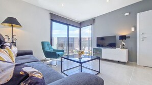 Een moderne woonkamer met een bank, een blauwgroene fauteuil, een glazen salontafel, een televisie en een balkon met uitzicht.