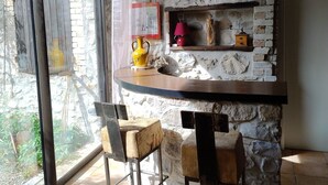 Une pièce aux murs de pierre, avec une table en bois et deux chaises, une lampe rouge et une petite étagère ornée d’objets décoratifs.