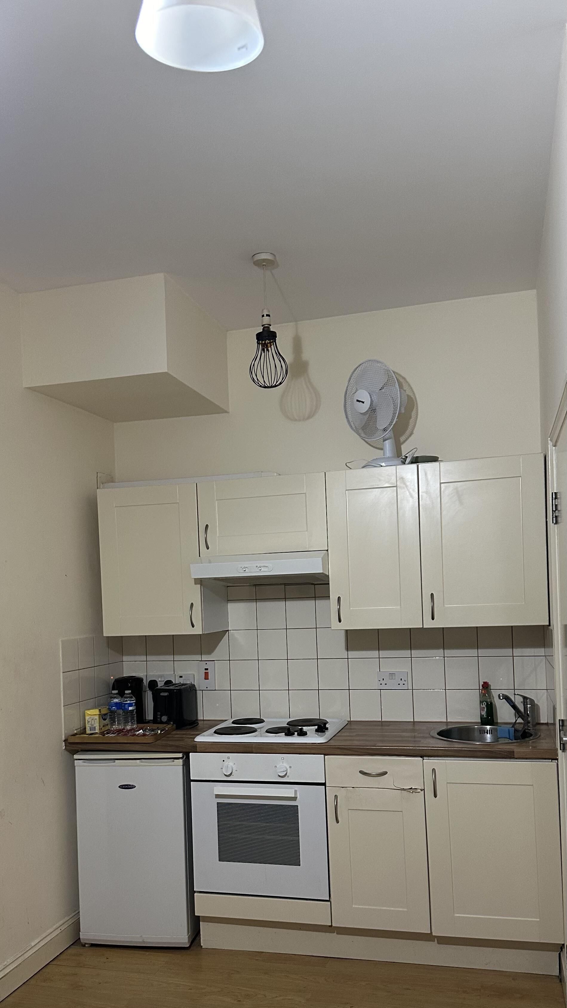 A compact kitchen with white cabinets, a small refrigerator, a stove, and a ceiling fan.