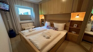 Een tweepersoonsbed met twee kussens en een deken, een nachtkastje, een lamp, een televisie en een raam met gordijnen.