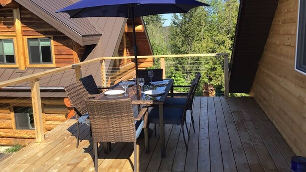 Een houten terras met een tafel, stoelen en een parasol, met uitzicht op een bebost gebied.