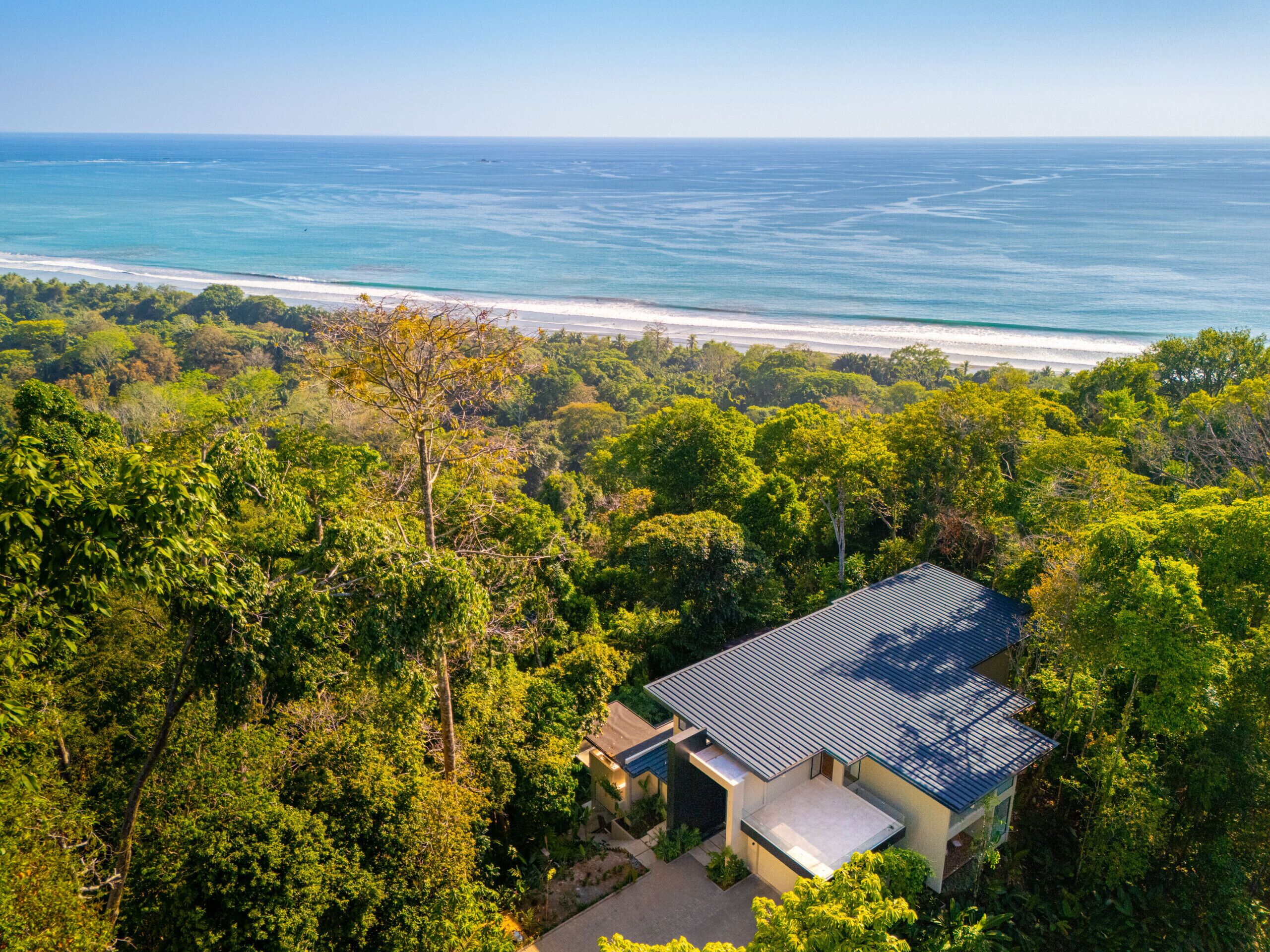 Vista aérea de una casa con techo metálico, rodeada de vegetación exuberante y con vistas a una playa de aguas azul cristalino.