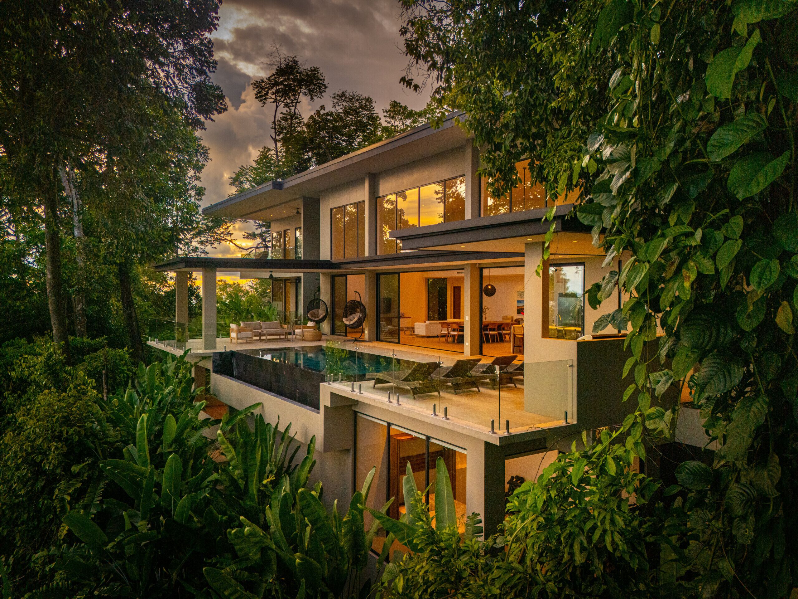 Una casa moderna de varios pisos con un amplio frente de vidrio, una piscina y abundante vegetación a su alrededor.
