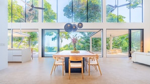 A spacious dining area with a large wooden table and chairs, large windows offering views of greenery, and a ceiling fan. - Casa Colibri by Pura Villas (Uvita)
