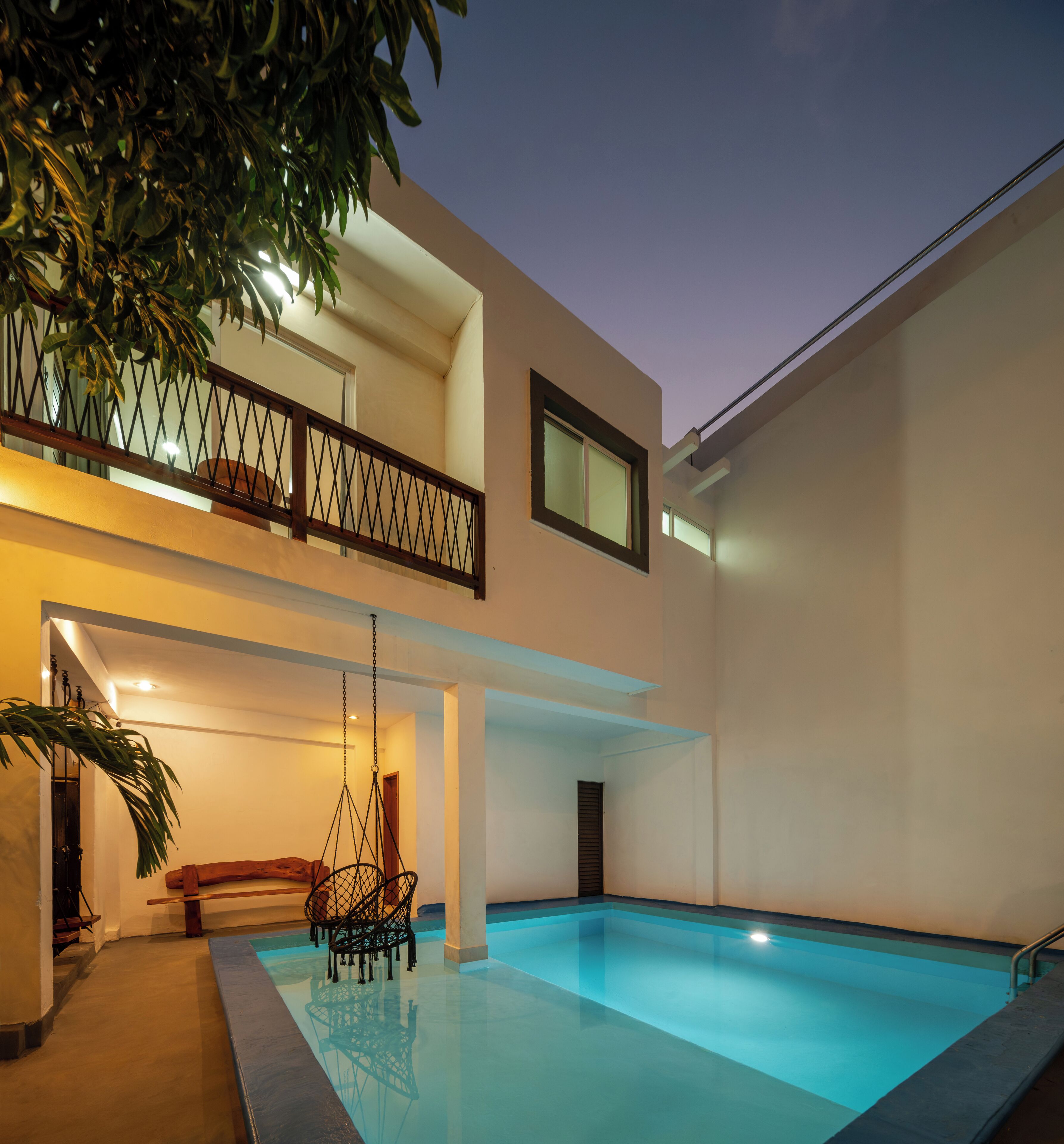 Una piscina interior moderna con techo de cristal, terraza de madera y una silla colgante.