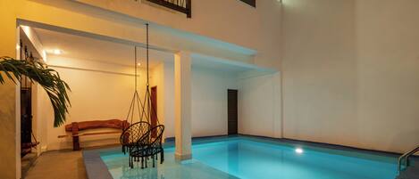 Una piscina interior moderna con techo de cristal, terraza de madera y una silla colgante.
