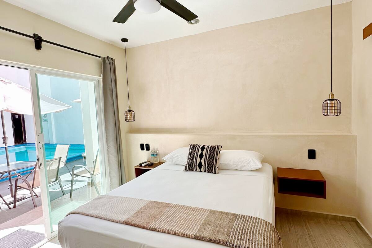 Quarto de hotel com cama, ventoinha no teto, mesas de cabeceira e vista para a área da piscina.