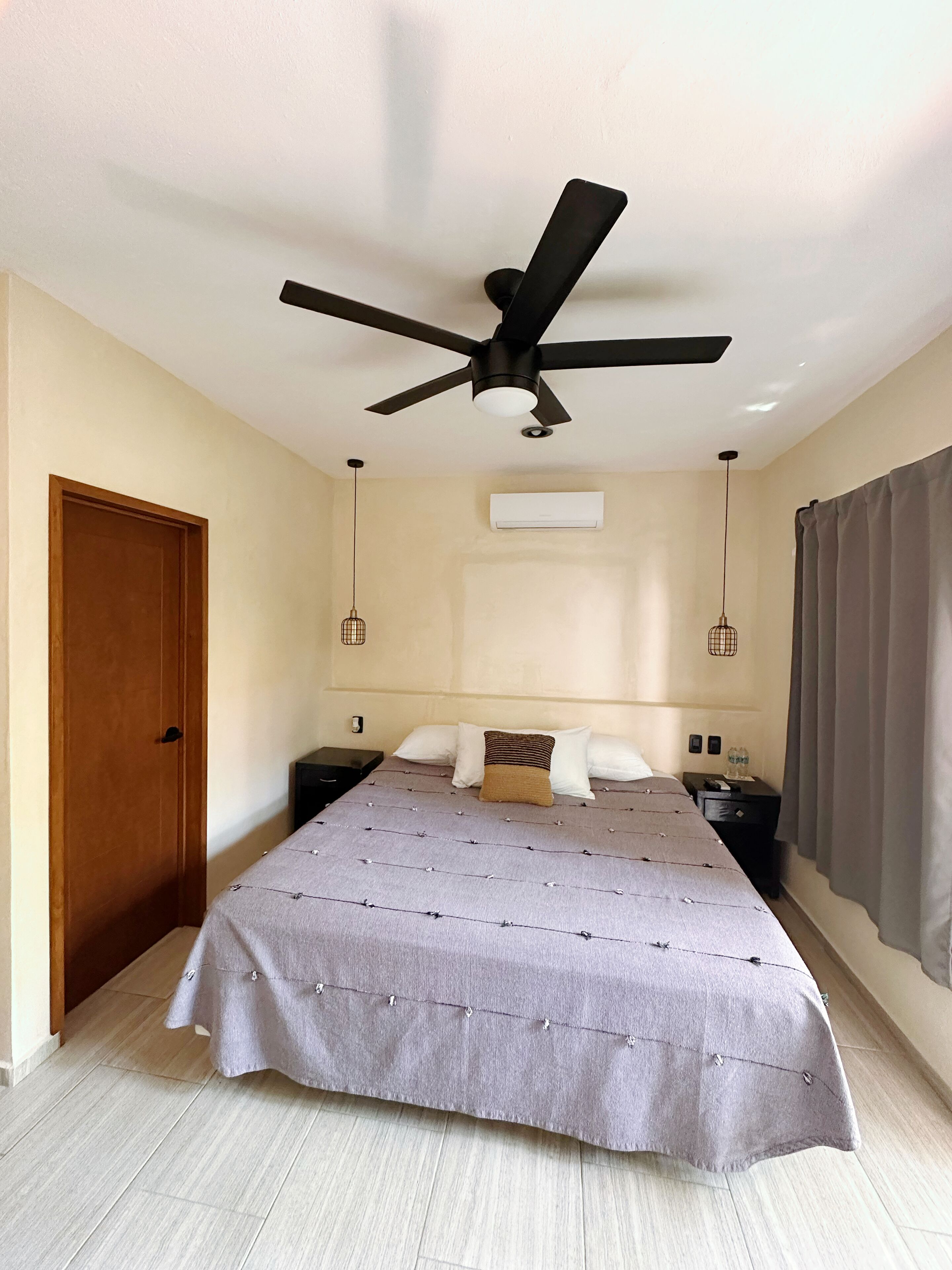 Une chambre à coucher comprenant un lit, un ventilateur de plafond, un appareil de climatisation, une table de chevet et une porte.