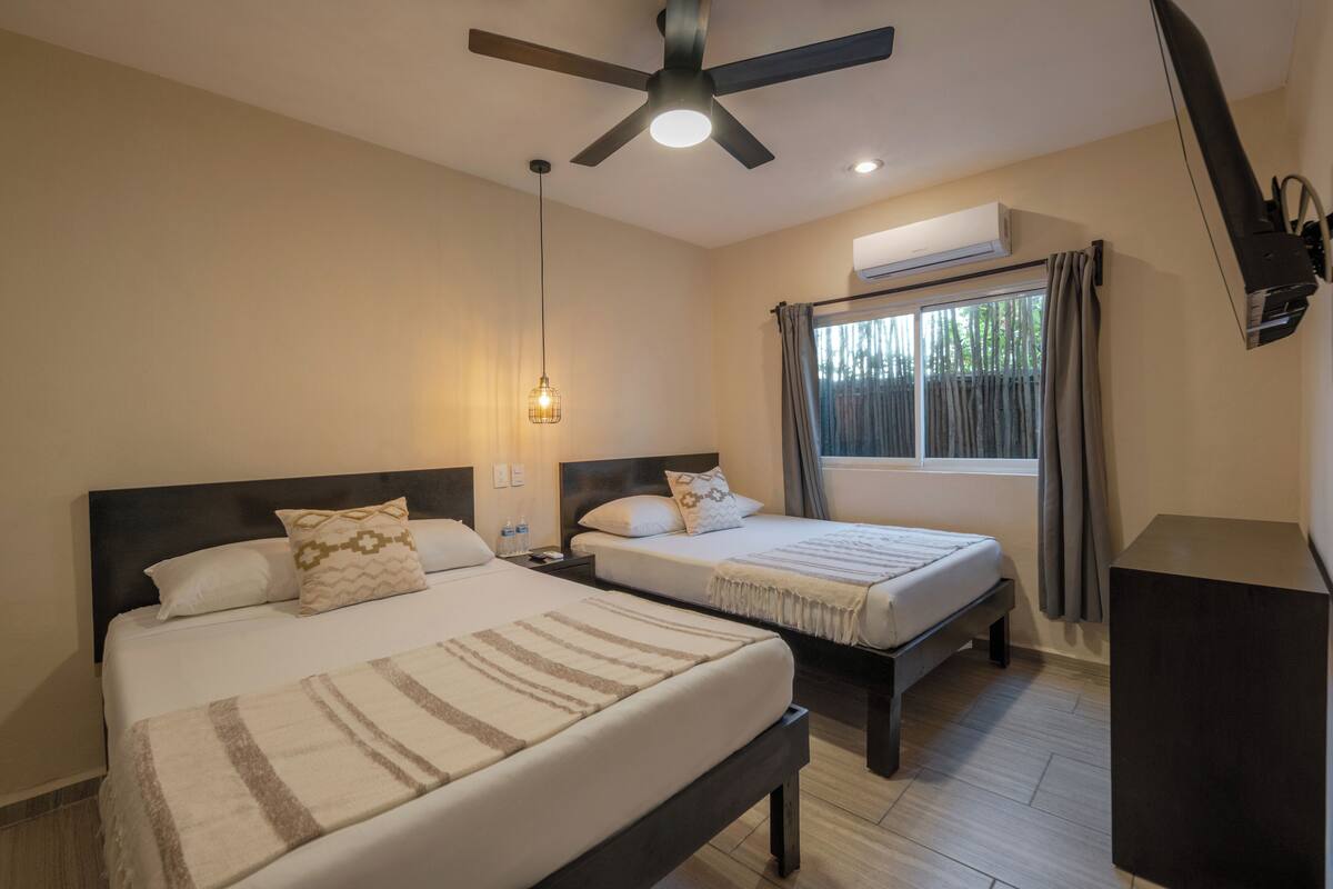 Quarto de hotel com duas camas, um ventilador de teto, uma janela com cortinas e uma mesinha de cabeceira.