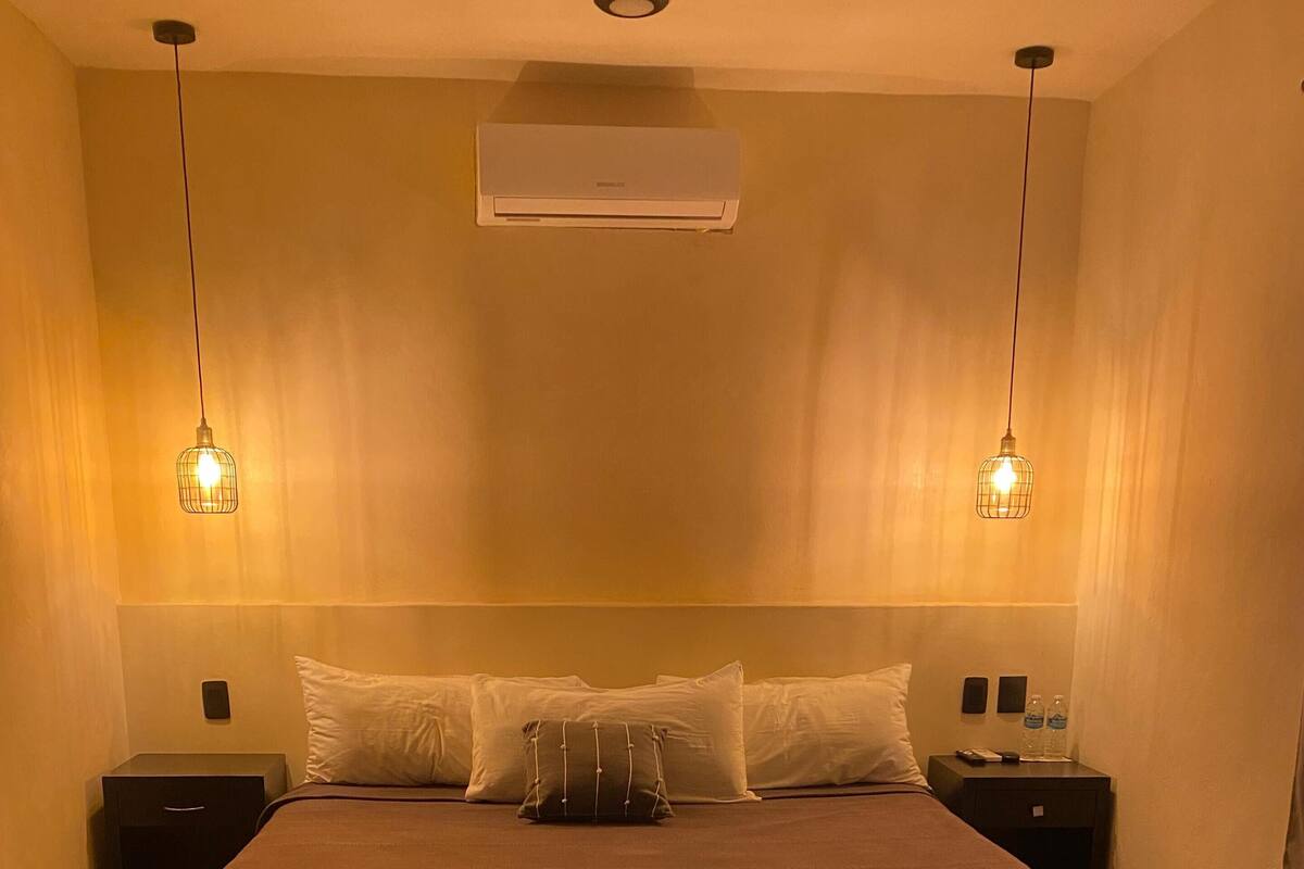 Uma cama bem arrumada com um edredom escuro, duas mesas de cabeceira com luminárias e um aparelho de ar condicionado acima da cama.