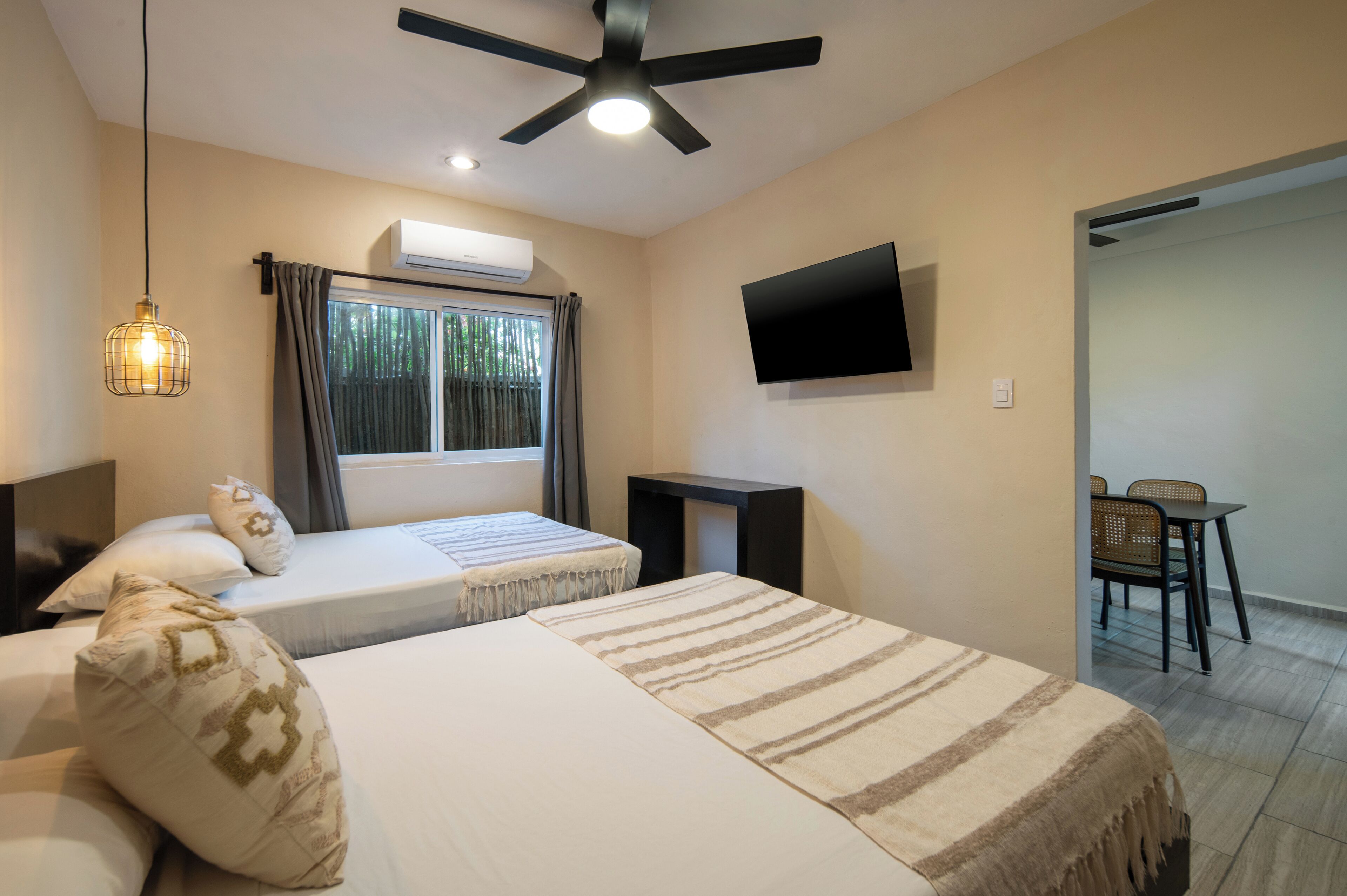 A hotel room with two beds, a ceiling fan, an air conditioner, a TV, a window with curtains, and a dining area with a table and chairs.