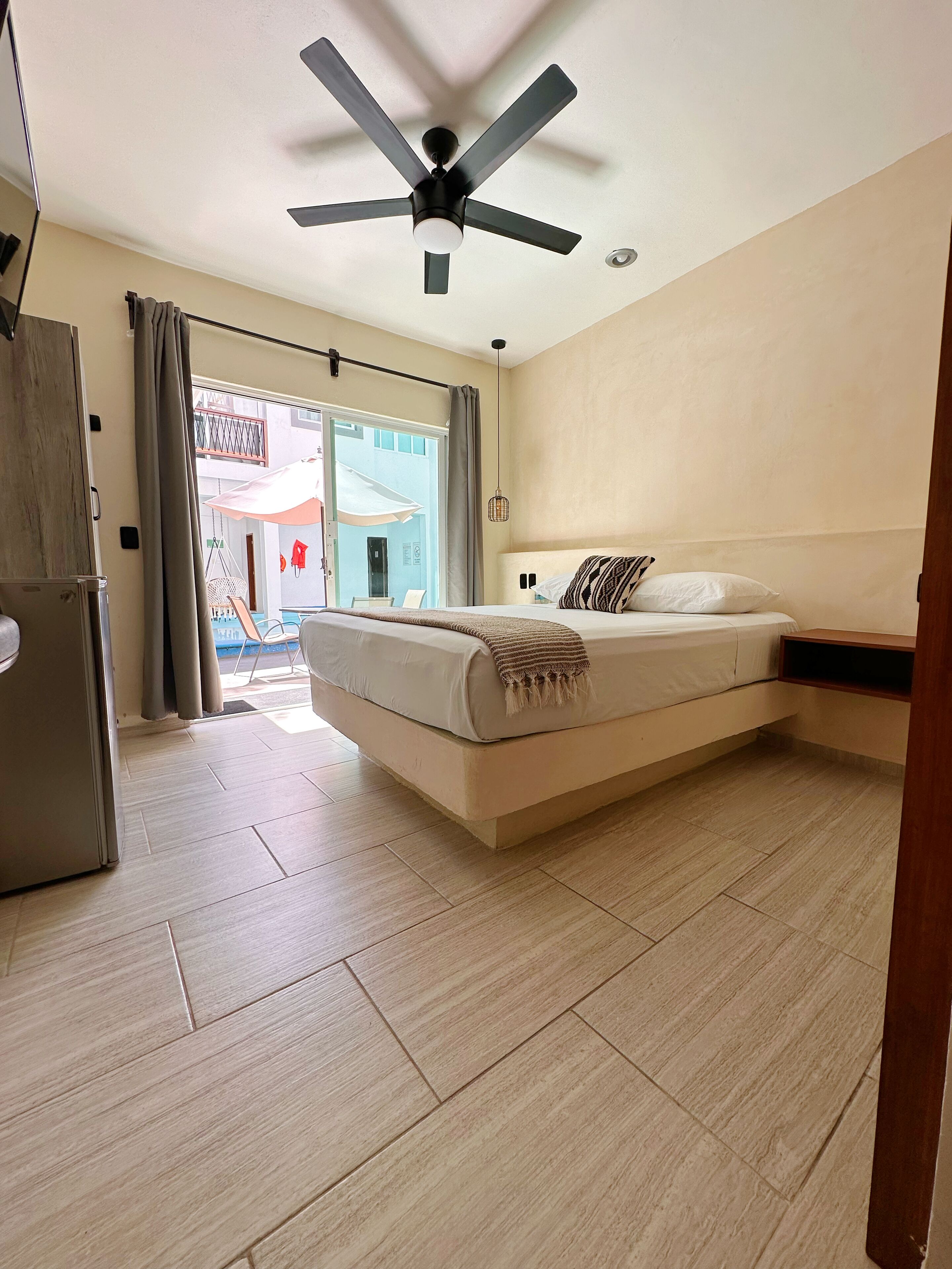 A bedroom with a bed, a ceiling fan, a refrigerator, and a view of the outside.