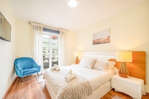 A hotel room with a bed, a blue chair, a bedside table, and a view of balconies through a window. - GuestReady - Happy Home in Santa Cruz (Lisbon)