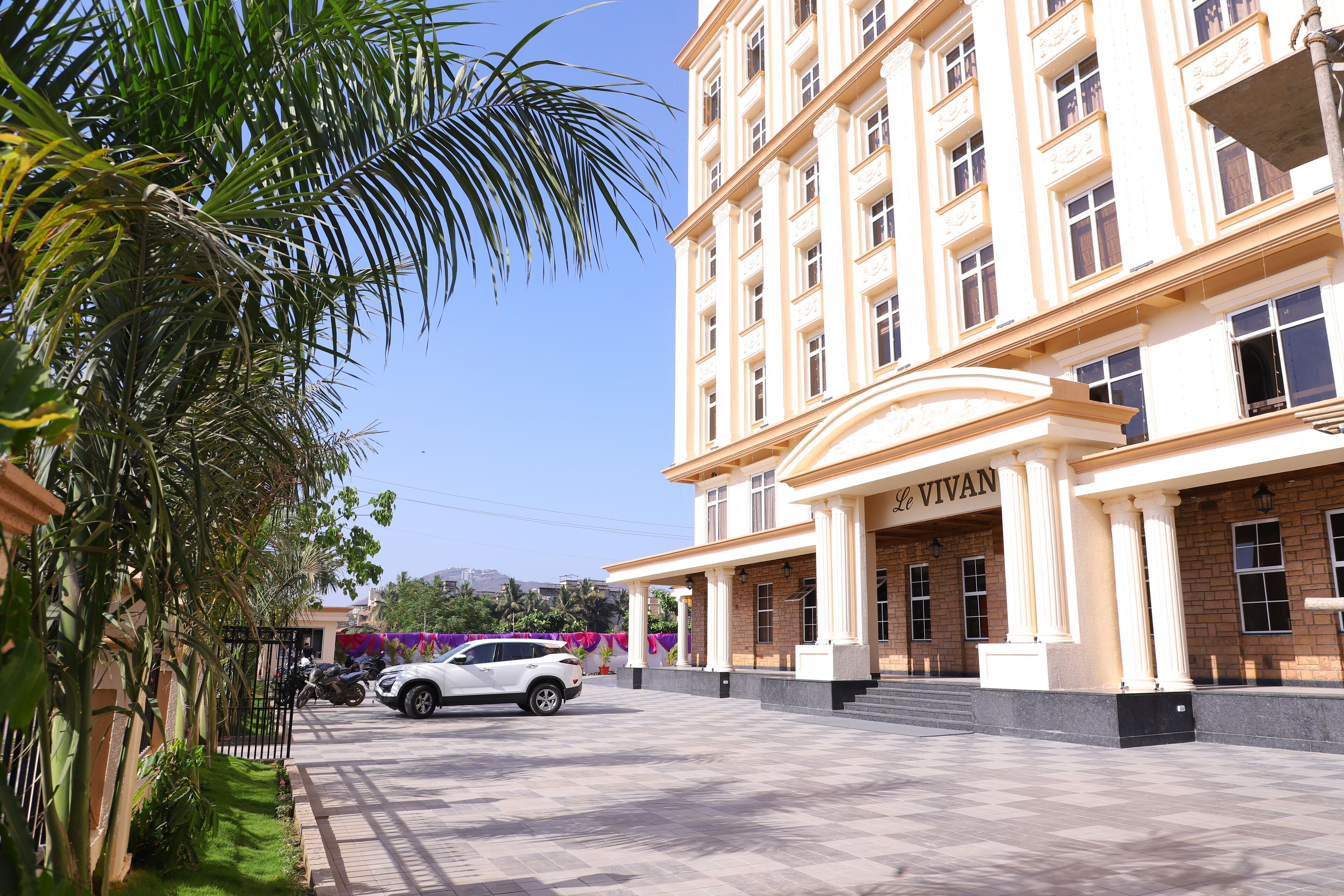 A hotel entrance with a white car parked in front, palm trees, and a clear blue sky.
