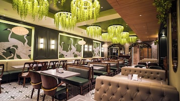 Interior de restaurante com paredes verdes, arte nas paredes em branco, plantas pendentes e diferentes opções de assento, incluindo boxes e mesas.