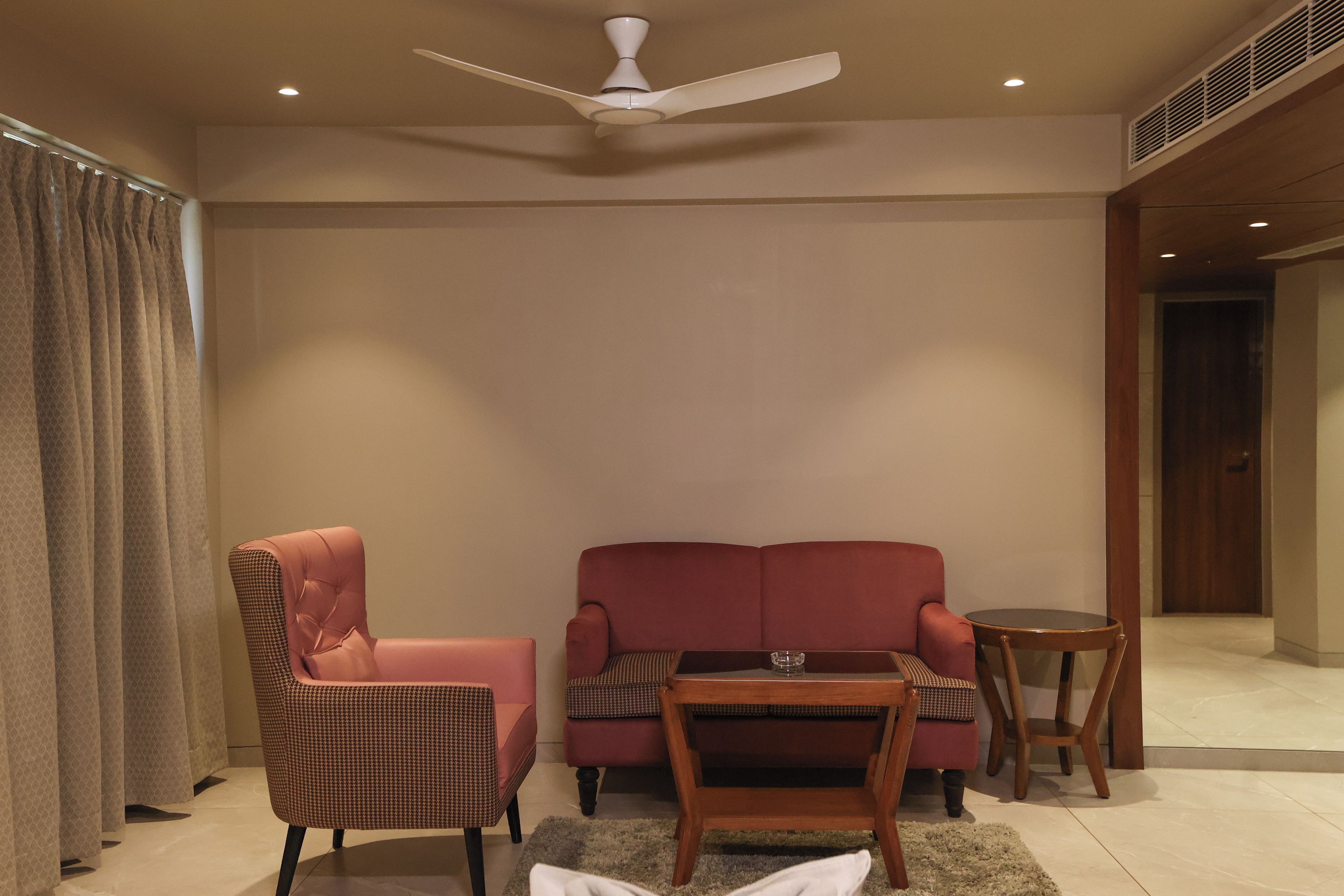 A room with a ceiling fan, a red sofa, a houndstooth armchair, a small round table with a glass, and a wooden chair.