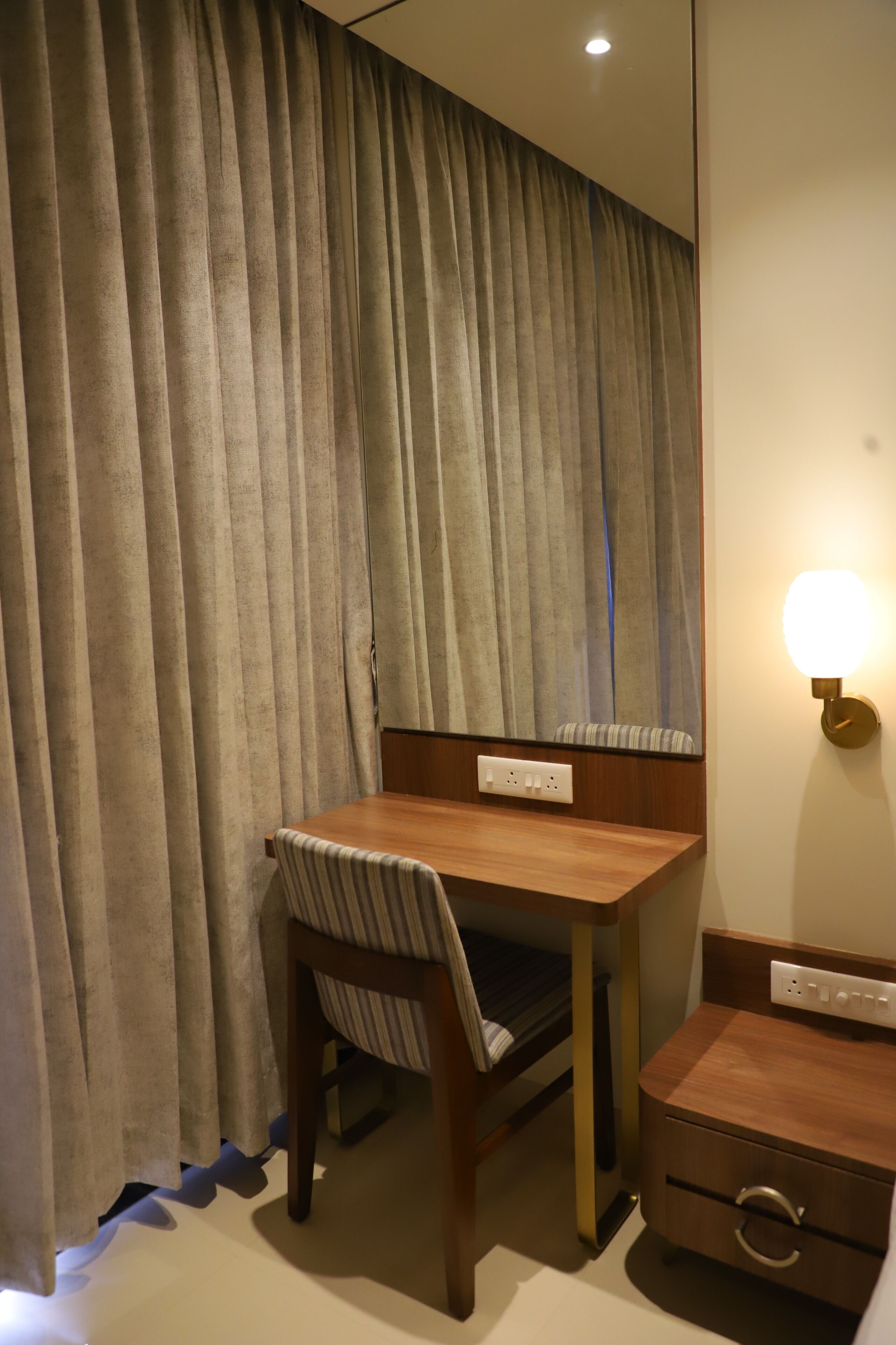 A hotel room with a wooden desk, a chair, a bedside table, and a wall-mounted light fixture.