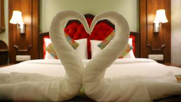 Una cama con cabecera roja y almohadas blancas, dos lámparas a cada lado, y un arreglo de toallas con forma de corazón sobre la cama.