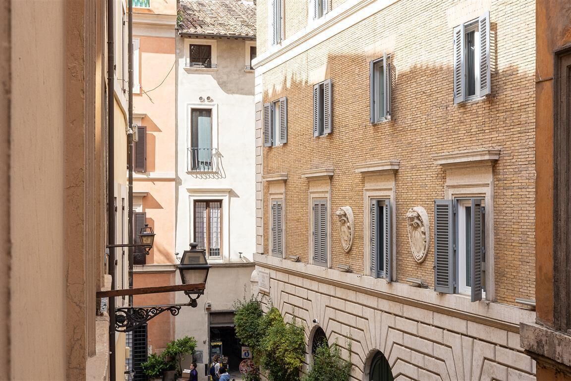 A narrow alleyway between buildings with traditional architecture, including a balcony with a wrought iron railing and a stone wall with a decorative shield.