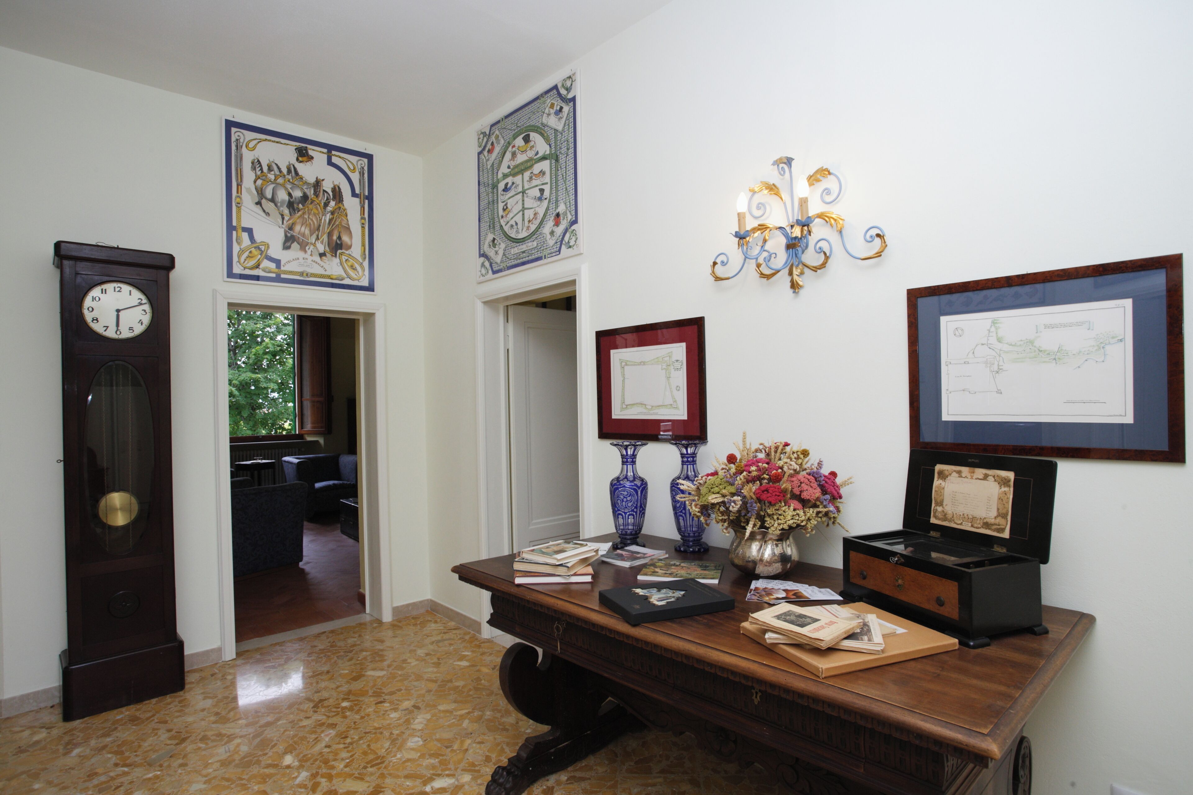 A hallway with a wooden desk, framed artwork, a clock, and a vase with flowers.