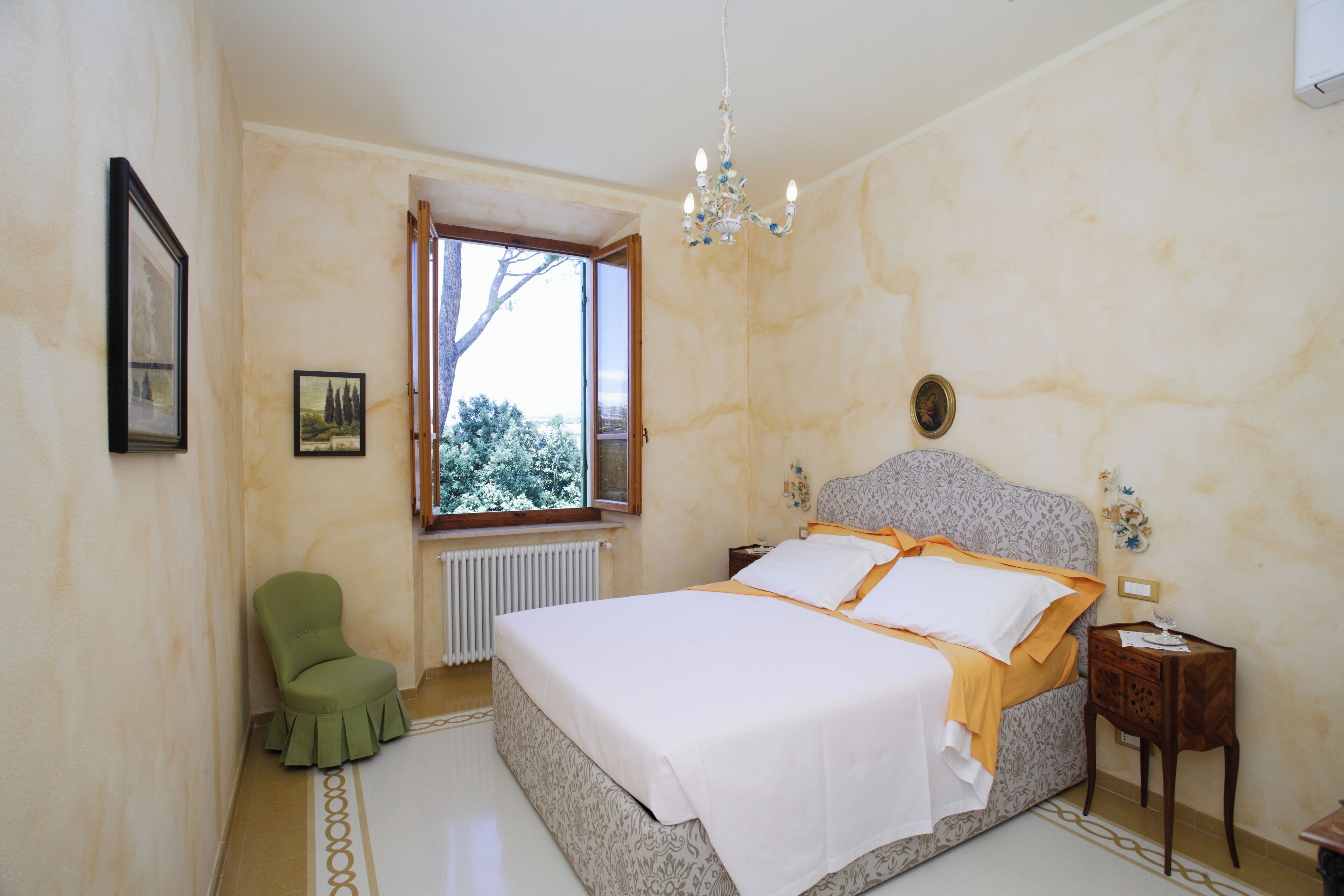A bedroom with a bed, a chair, a nightstand, a chandelier, and a window with a view of trees.