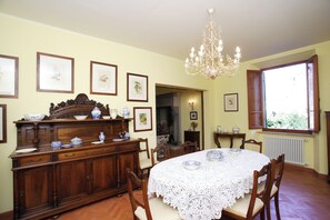 Ein Esszimmer mit einem runden Tisch, der mit einer Spitzentischdecke bedeckt ist, Holzstühlen, einer Holzkommode und gerahmten Bildern an den Wänden.