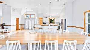 Un vaste espace de salle à manger avec une longue table en bois et des chaises blanches, une cuisine équipée d’appareils électroménagers modernes et une grande fenêtre laissant entrer la lumière naturelle.