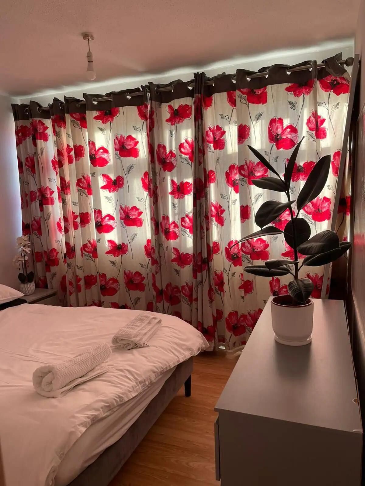 A bedroom with a bed, a bedside table, a potted plant, and curtains with a floral pattern.