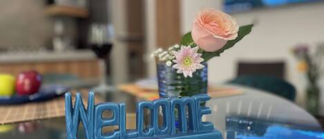Une plaque bleue avec l’inscription « Bienvenue » est posée sur une table en verre, à côté d’une rose et d’un petit vase rempli de fleurs.