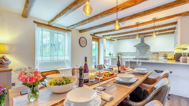 Un comedor con una mesa de madera preparada para una comida, una cocina al fondo y un reloj en la pared.