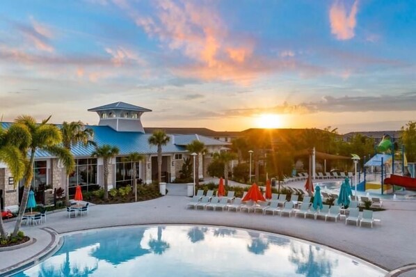 A resort pool area with lounge chairs and umbrellas, a building with a blue roof, and a sunset in the background.
