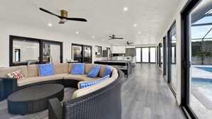 A modern living room with a curved sectional sofa, a dining area, and a kitchen. There are ceiling fans and large windows providing natural light. - Naples Oasis: Heated Pool, Gazebo, Grill Retreat (Naples)