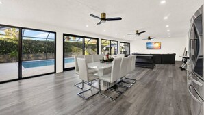 A modern kitchen with a large island, a dining area with a white table and chairs, and a pool visible through large windows. - Naples Oasis: Heated Pool, Gazebo, Grill Retreat (Naples)