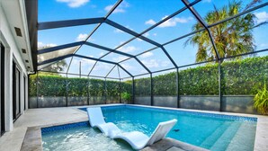 A covered swimming pool with two white lounge chairs, surrounded by a metal frame and glass walls, with a view of palm trees and a hedge in the background. - Naples Oasis: Heated Pool, Gazebo, Grill Retreat (Naples)