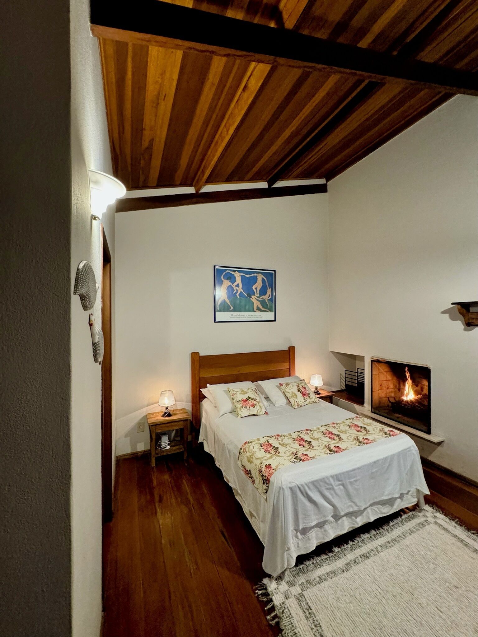 Un dormitorio con techo de madera, cama con ropa de cama floral, mesita de noche con lámpara y chimenea.