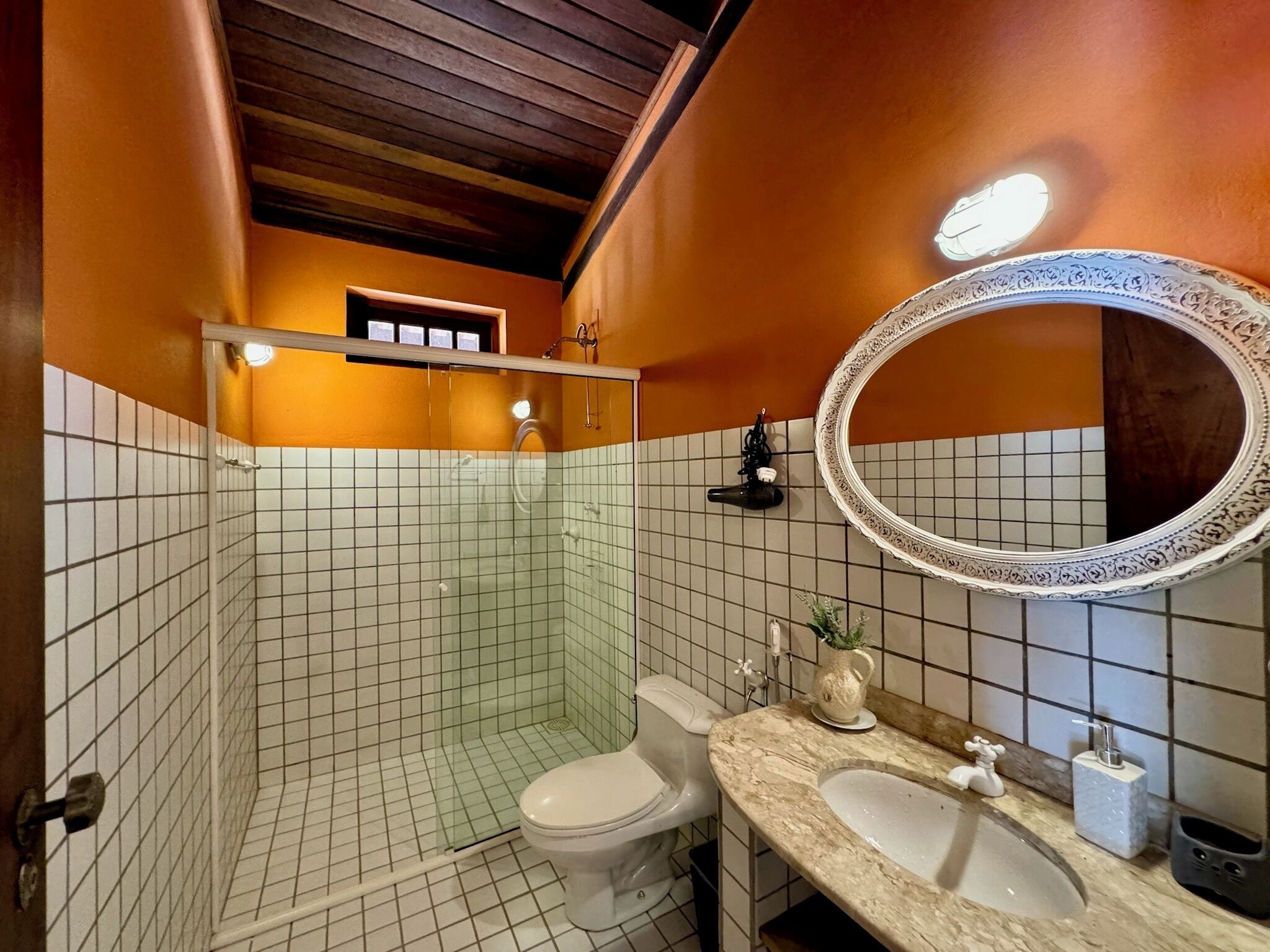 Baño con inodoro, lavamanos y zona de ducha, con azulejos blancos y techo de madera.