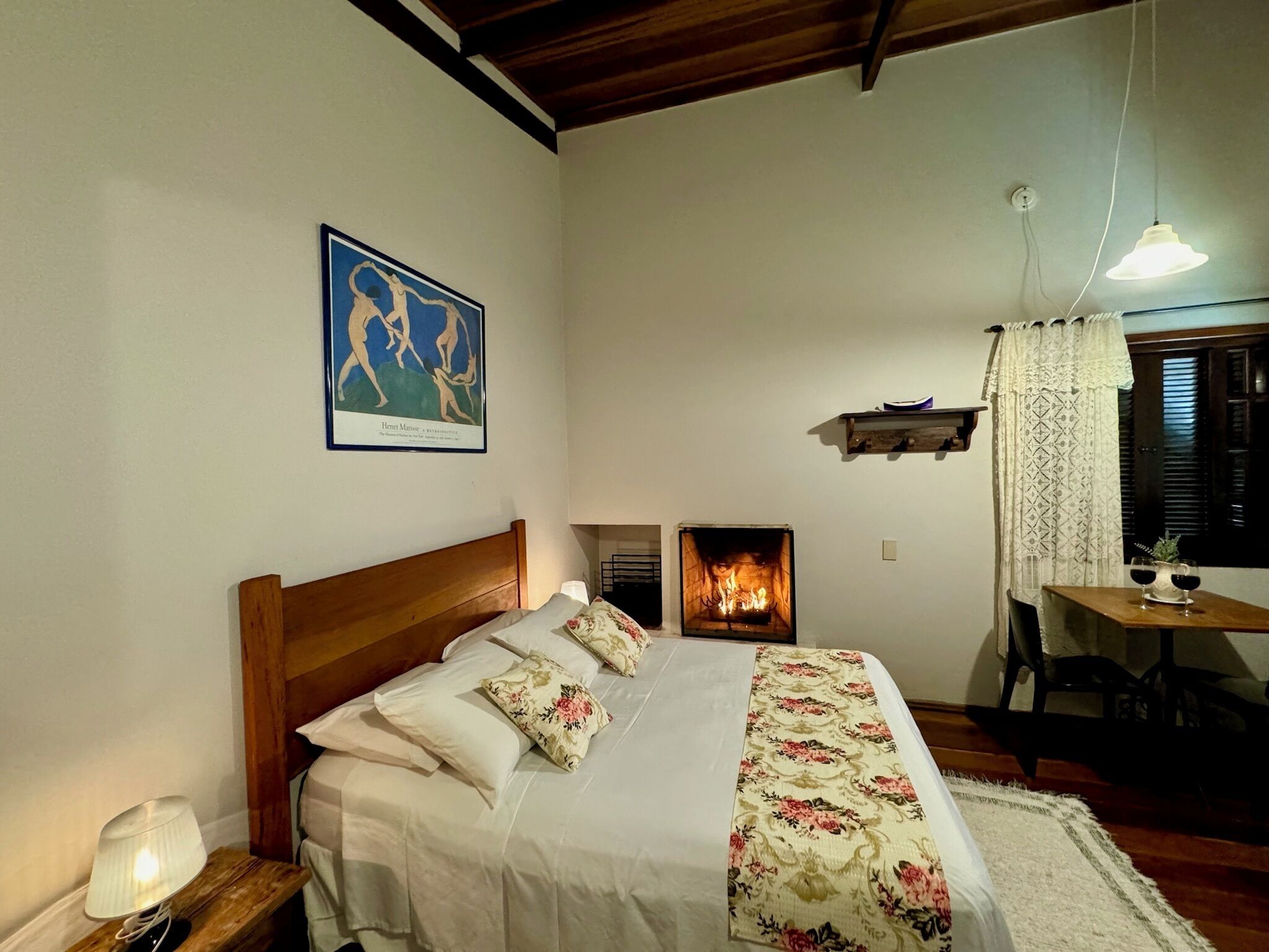 A bedroom with a wooden bed, a nightstand with a lamp, a fireplace, a table with two glasses of wine, and a window with curtains.