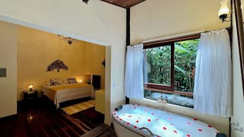A bathroom with a bathtub, a window with curtains, a bed with a nightstand, and a wall-mounted light fixture.