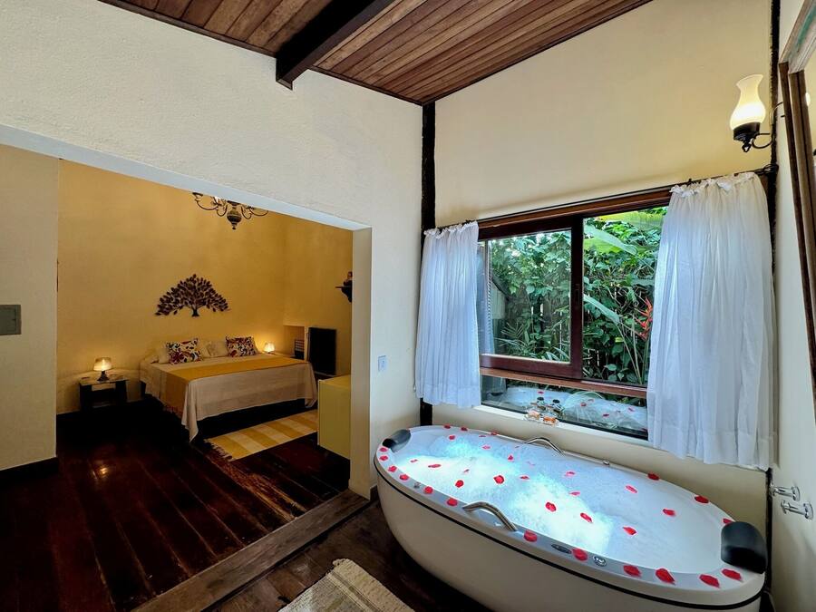A bathroom with a bathtub, a window with curtains, a bed with a nightstand, and a wall-mounted light fixture.